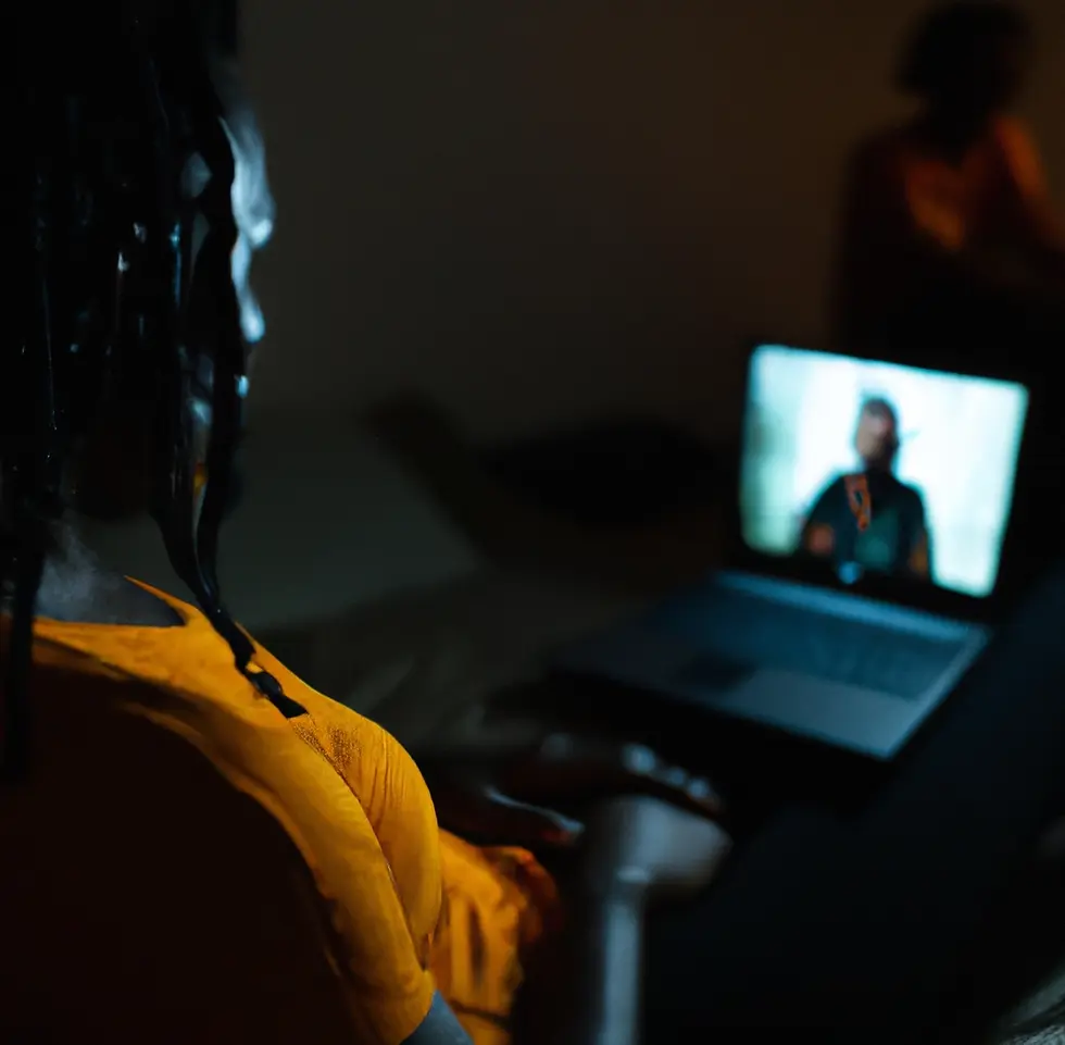 A person alone in a dark room wearing a yellow shirt watching a laptop screen with a man blurred out on it. The feeling is one of being alone but help and hope being available.