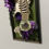 Thumbnail: A clear glass diffuser jar filled with Lemon scented fragrance oil meant for hanging from a cars rear view mirror. In the background there is a picture frame with a skeletons rib cage and purple flowers.  