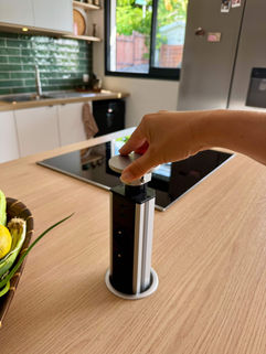 Main qui déploie une prise escamotable dans un îlot de cuisine en bois clair sur-mesure.