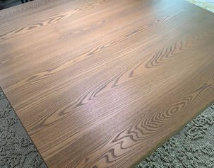An overhead view focusing on the rich, flowing grain pattern of a wide, light-brown stained wood table surface.