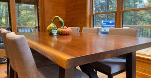 A polished, thick wooden dining table with a glossy reflective top and dark legs, surrounded by matching chairs in front of large windows.