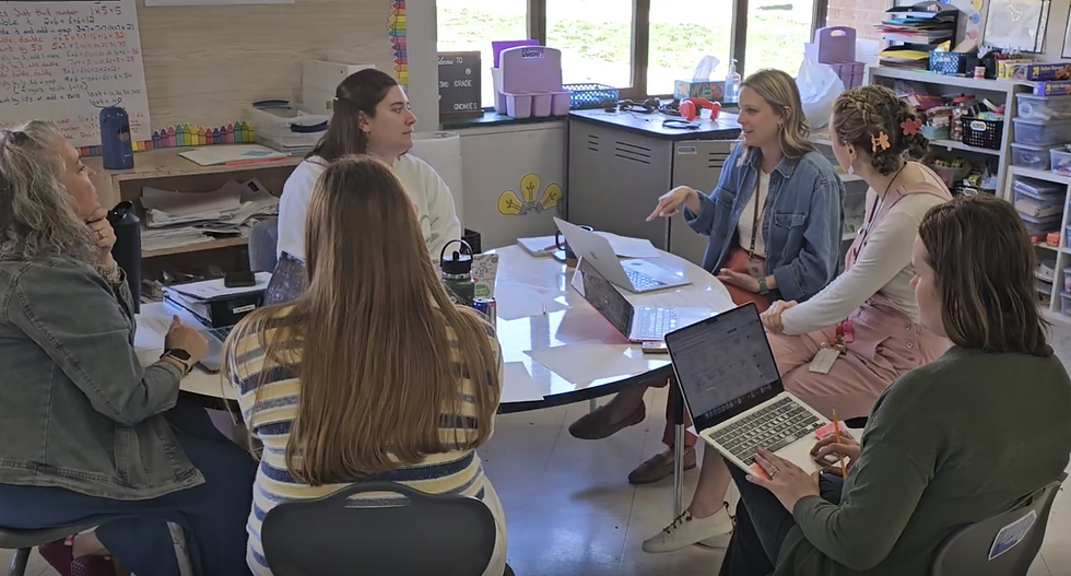 A PBL coach meets with a team of teachers around a classroom table, collaborating on project plans using laptops and shared materials.