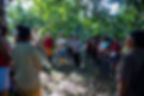 A group of people gather in a lush, green forest, engaged in conversation. Sunlight filters through the leaves, creating a serene atmosphere.