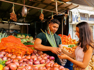 Market scene with a vendor handing vegetables to a woman. Colorful produce surrounds them. Both appear engaged in conversation.