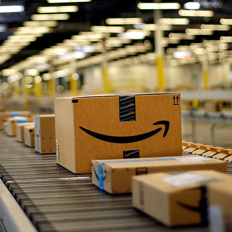 Caixas de papelão da Amazon em esteira transportadora em armazém iluminado. Ambiente industrial com foco no logotipo preto da caixa.