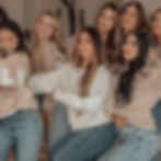 A group of smiling women in beige sweaters and jeans pose cheerfully together indoors, with a cactus in the background.