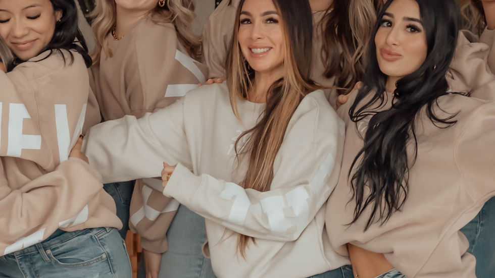 Group of women smiling, wearing beige sweatshirts and jeans, posing together indoors with a casual vibe. Cactus visible in background.