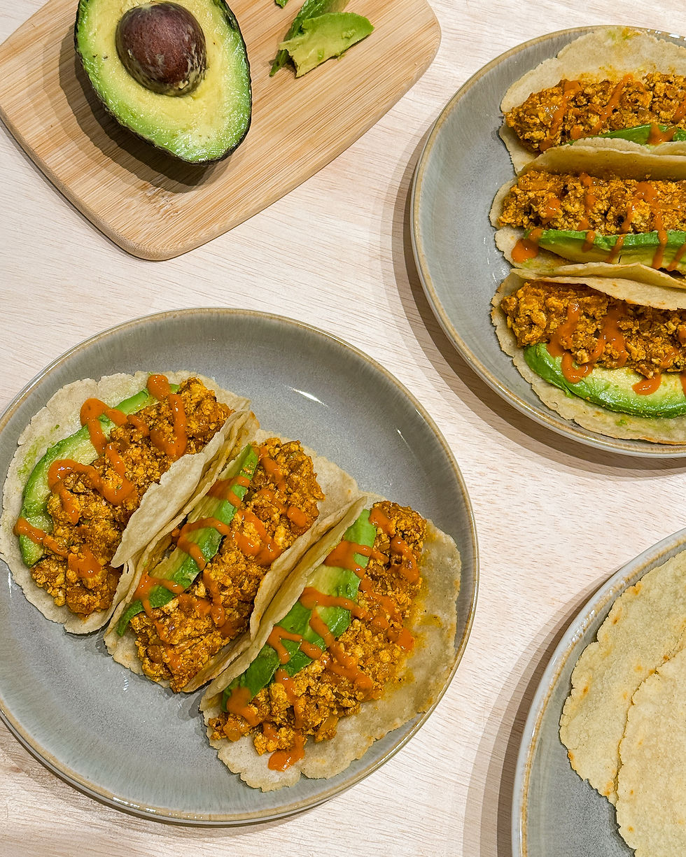 Three tacos with tofu, avocado, and hot sauce on a gray plate. A halved avocado on a wooden board. Neutral wooden table background.
