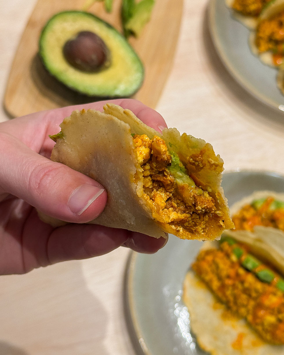 Hand holding a taco filled with tofu and avocado, with a sliced avocado on a wooden board in the background. Earthy tones, casual setting.