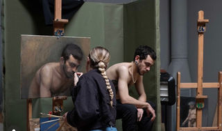 Woman painting from a live model with the man facing down. Side view.