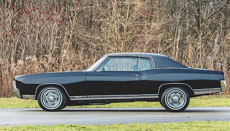 Black classic car parked on a road beside leafless trees and grass. Silver trim and chrome wheels stand out in the natural setting.