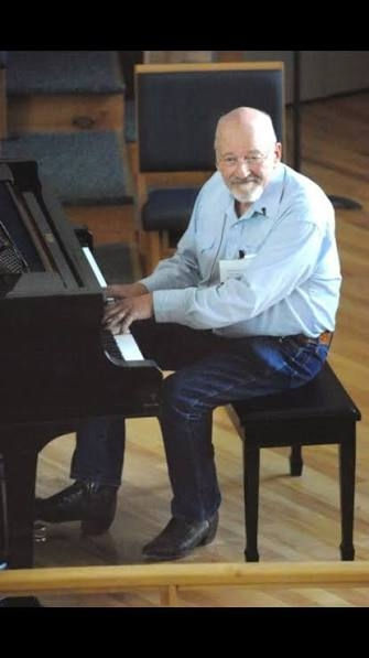 Elderly man in a light blue shirt playing a black piano, smiling warmly in a spacious room with wood floors and chairs in the background.