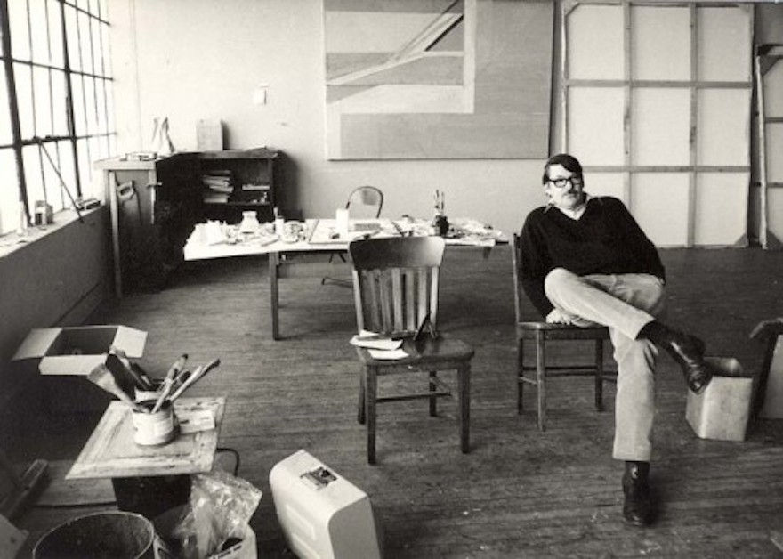 Man sitting casually in a chair in a cluttered studio with large windows and abstract art on walls. Monochrome, relaxed atmosphere.
