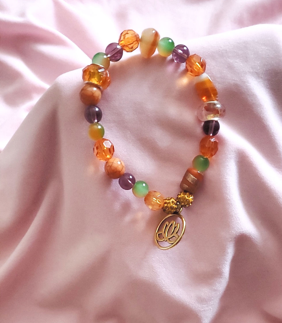 A vibrant, bohemian-style stretch bracelet with a mix of orange, purple, and green faceted and glass beads, featuring a dangling gold lotus flower charm, on a soft pink fabric.