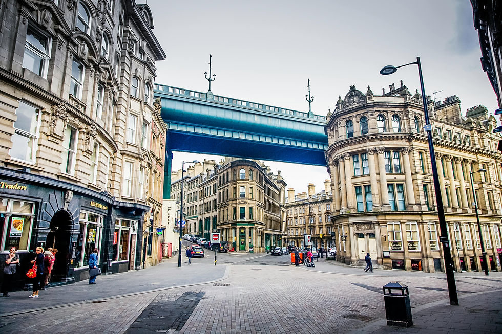 Street view of Newcastle city with historic buildings, a the Tyne bridge overhead, and people walking. Overcast sky adds a subdued mood.