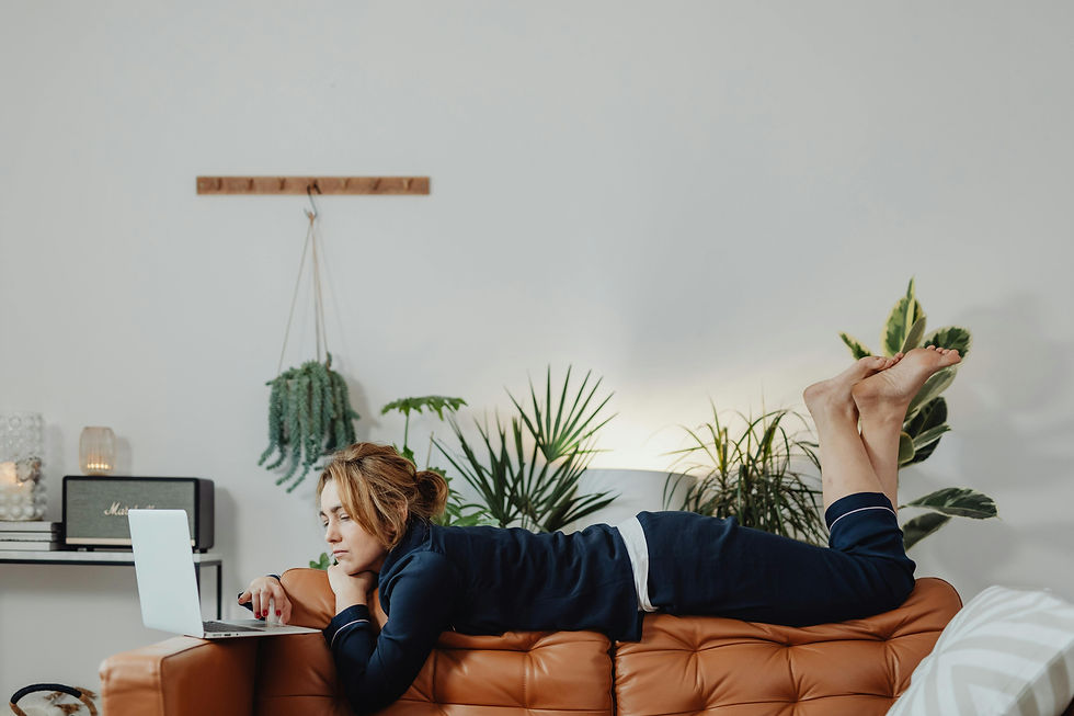 Woman in navy loungewear lies on a tan couch using a laptop, in a cozy plant-filled living room.