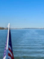 American flag waving on a boat, a plane flying overhead, and cityscape in the distance