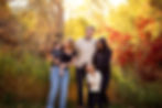 A family of four smiles in a sunlit garden with autumn colors. The mother holds a toddler, while the father and older child share a moment. Photographed by Tiffany Edwards family photographer at Riverfront Park in Billings MT