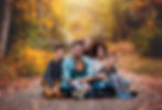 Family of six sits on a leaf-strewn path in an autumn forest, smiling warmly. Bright fall colors surround them, creating a cozy atmosphere as photographed at Riverfront Park by Tiffany Edwards, family photographer