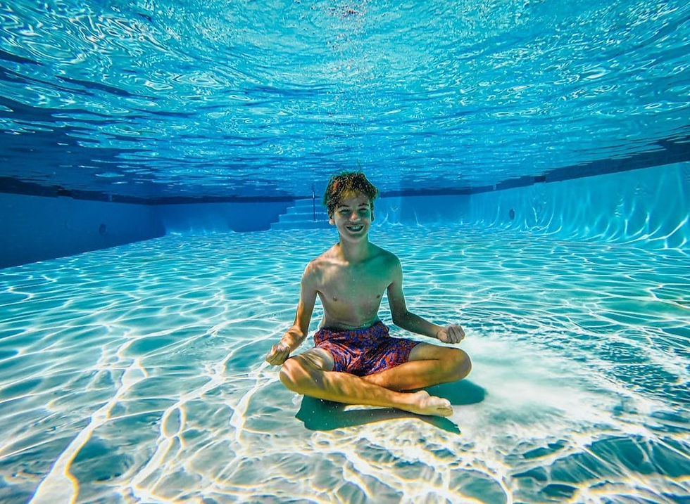 Underwater view of a smiling boy sitting cross-legged on the floor of a bright, sunlit swimming pool, demonstrating the "floor sit" technique with sunlight rippling across the pool bottom.