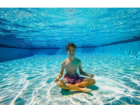 Underwater view of a smiling boy sitting cross-legged on the floor of a bright, sunlit swimming pool, demonstrating the "floor sit" technique with sunlight rippling across the pool bottom.