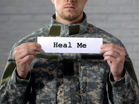 Person in camouflage uniform holding a sign reading "Heal Me" against a gray brick wall, conveying a plea or emotional appeal.