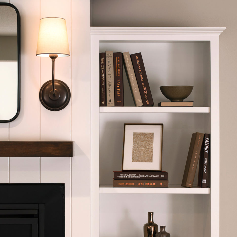 A vignette of a fireplace surround features clean white painted custom cabinetry and tongue and groove planks. The open bookshelves are styled with neutral books,  small frame art, and ceramics. A simple bronze sconce is installed above a floating walnut mantle, next to a modern black framed mirror.