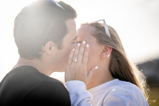 Close-up of a couple sharing a kiss outdoors, with the engagement ring visible as the bride-to-be holds her hand against her partner’s face.