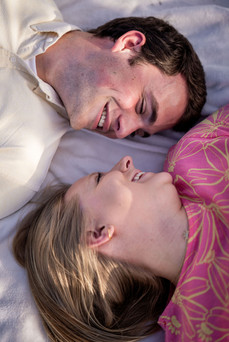 Romantic engagement portrait of a couple lying side by side on a blanket, gazing at each other with loving smiles.