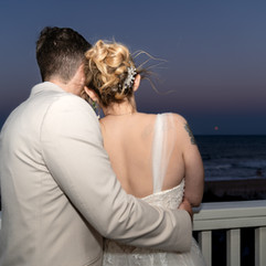 Bride and groom embracing on balcony overlooking ocean at sunset wedding