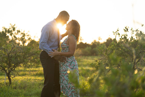 Romantic engagement photo of a couple standing close in a field at sunset, sharing an intimate moment with golden light behind them.