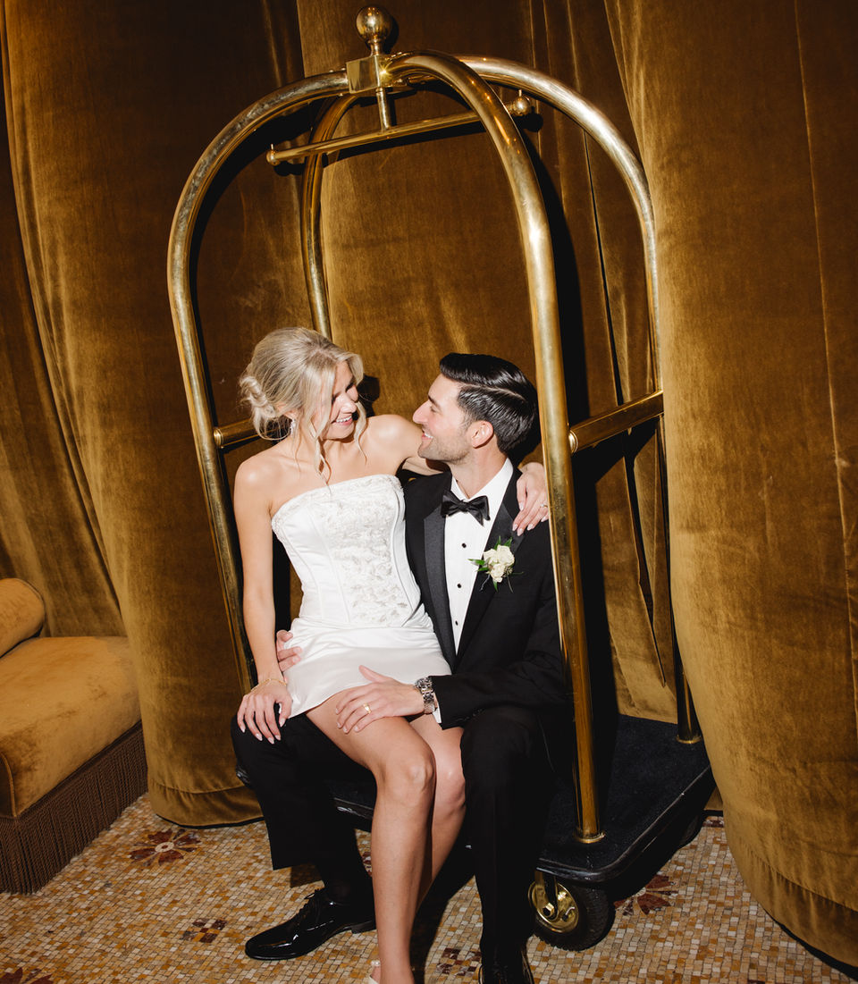 Modern bride and groom sitting on a luggage cart in a hotel lobby on their wedding day.