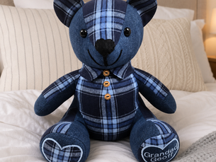 A handmade memory bear from a loved one's clothing by Deborah Jameson Creations, resting on a bed in soft light
