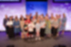 Group photo of Festival of Quilts 2024 award winners on stage, featuring quilters of all ages and backgrounds smiling in front of the event backdrop