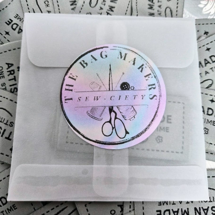 Branded envelope from The Bag Makers Sew-Ciety resting on top of artisan woven labels with a soft grey and black design.