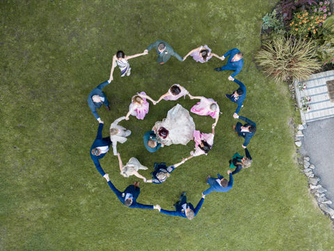 Wedding party holds hands in a circle on grass, aerial view. Mo Photography.