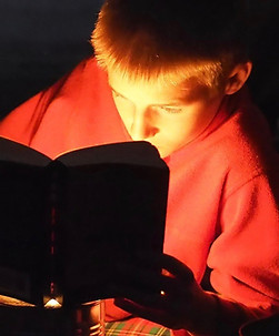 A boy reads a book by candlelight next to a campfire in his tipi