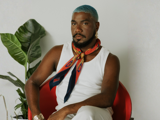 O homem negro elegante com cabelo curto azul vibrante e barba está sentado em uma cadeira de design preta. Ele veste uma regata branca e um lenço de seda estampado colorido (tons de laranja, azul, vermelho e dourado) amarrado no pescoço. Ele olha confiantemente para a câmera, usando um brinco de argola dourado e uma pulseira dourada larga no pulso. Uma planta grande em um vaso branco está à esquerda no fundo neutro.