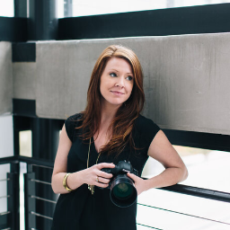 Woman with long hair, holding a camera, leans on an indoor railing. She wears a black top, looks thoughtful. Concrete and metal decor.