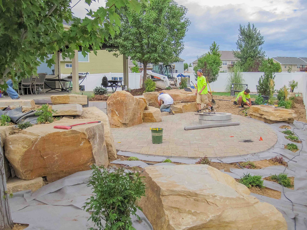 Workers installing a stone patio and garden in a suburban backyard, with boulders, plants, a skid steer, and overcast skies.