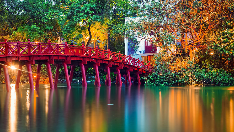 Red bridge over calm water, surrounded by lush trees with warm lights reflecting in the evening. Peaceful and colorful setting.