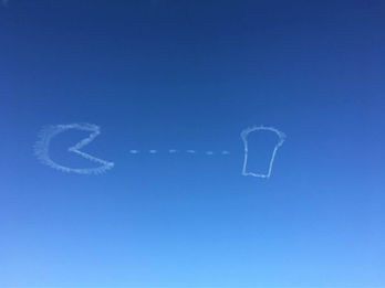 Pac-Man and ghost symbol skywriting message with trail of dots in a clear blue sky, created in collaboration with The Skywriters, showcasing precision skytyping, skywriting, aerial advertising, and high-impact event marketing and wide area engagement opportunities.