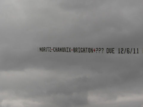 Sky banner featuring the message "MORITZ-CHAMONIX-BRIGHTON+??? DUE 12/6/11" in a mix of black and red block letters, flying against a dark, overcast sky, created in collaboration with The Skywriters, showcasing precision aerial advertising, creative gender reveal services and aerial sky banner messages.