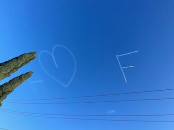 H heart F skywriting message in flowing cloud letters and a heart symbol in a clear blue sky above cypress trees and utility lines, created in collaboration with The Skywriters, showcasing precision skytyping, skywriting, aerial messaging, creative proposal ideas, and thoughtful messaging services.