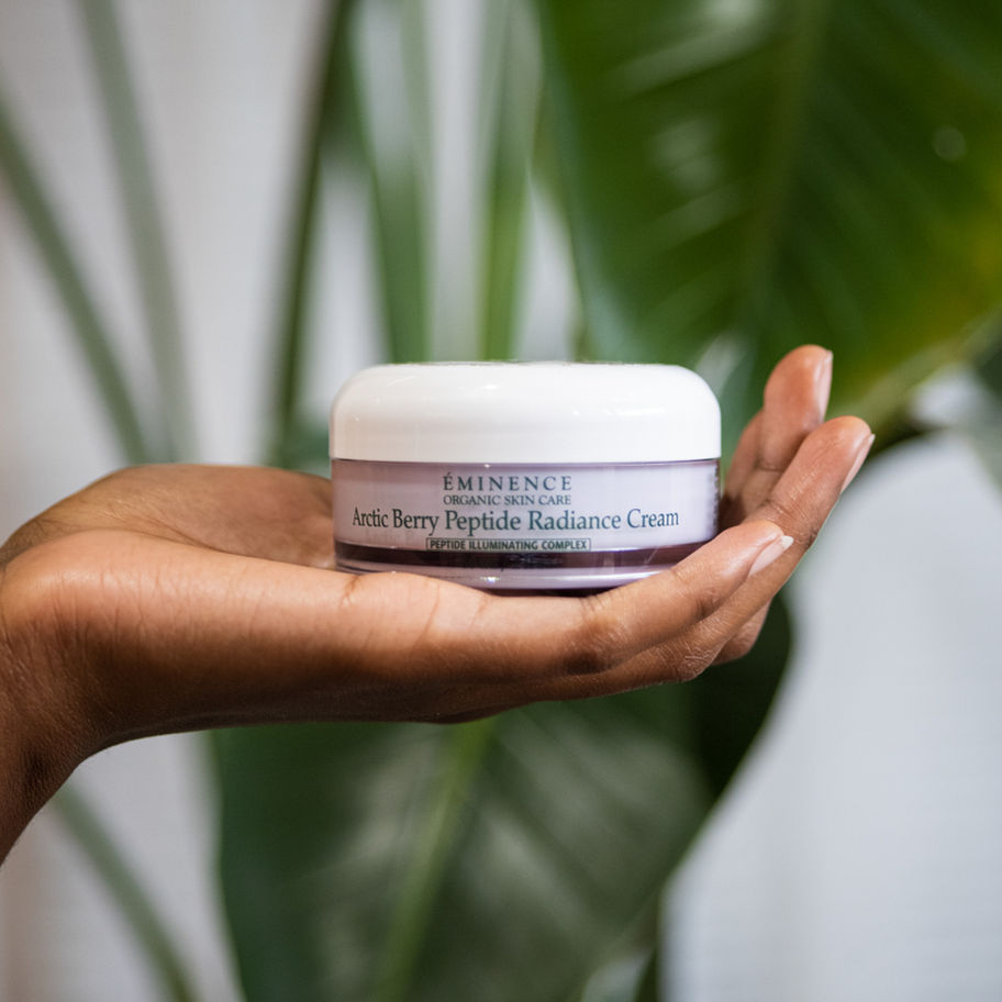 Hand holding jar of Acai Berry Peptide Radiance Cream, with green leaves background.