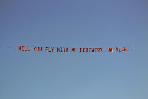 Custom airplane banner for a marriage proposal event announcing Will you fly with me forever? Alan, flying against a blue sky, created in collaboration with The Skywriters, showcasing professional aerial banner advertising, unique marriage proposal ideas, and high-impact aerial messaging for romantic celebrations.
