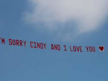 Sky banner featuring the message "I'M SORRY CINDY AND I LOVE YOU heart" in red block letters, flying against a blue sky with soft white clouds, created in collaboration with The Skywriters, highlighting thoughtful aerial messaging, creative sky banner moments,  and high-impact event marketing.
