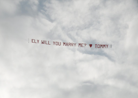 Custom airplane banner for a marriage proposal event announcing Ely Will You Marry Me? followed by a red heart icon and the name Tommy, flying against a cloudy white sky, created in collaboration with The Skywriters, showcasing professional aerial banner advertising, unique marriage proposal ideas, and high-impact aerial messaging for romantic celebrations.
