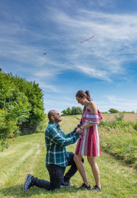 Marriage proposal event for a couple in a grassy field with a man on one knee holding an engagement ring box, while an airplane banner with a red proposal message flies in the distance against a bright blue sky, created in collaboration with The Skywriters, showcasing professional aerial banner advertising, unique marriage proposal ideas, and high-impact aerial messaging for romantic celebrations.
