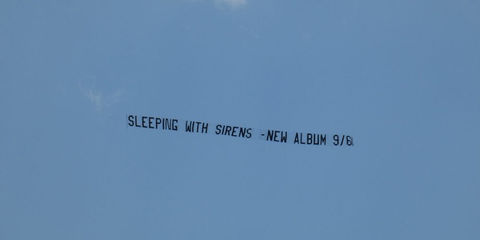 Sky banner featuring the message "SLEEPING WITH SIRENS - NEW ALBUM 9/6" in black block letters, flying against a clear blue sky, created in collaboration with The Skywriters to promote the band's latest release, showcasing precision aerial advertising and high-impact event marketing.